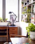 Home office with wooden desk, chair, desk lamp, potted plants, books, and framed art near a large window.
