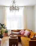 Living room with mustard sectional sofa, colorful pillows, wooden sideboard, potted plants, and a chandelier.