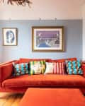Orange sofa with colorful patterned cushions, framed art on blue walls, and wooden floor in a cozy living room.