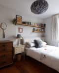 A floating bookshelf full of books and art above a simple white twin bed