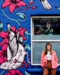 Woman in front of a vibrant mural with pink flowers and white birds on a blue wall.