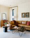 Living room with a tan leather sofa, colorful cushions, coffee table, art, and large windows with white curtains.