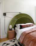A pink velvety comforter on a bed with an arched green headboard and a modern sconce