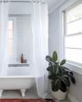 Clawfoot bathtub with white curtain, geometric floor tiles, potted plant, and abstract wall art in a bright bathroom.
