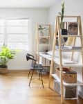 Home office with dual monitors on a wooden desk, ergonomic chair, ladder shelves, and potted plants by a window.