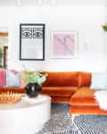 Orange velvet sectional with colorful pillows, abstract wall art, hanging plants, and a chess set on a round table.
