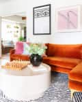 Living room with orange velvet sofa, round coffee table, chess set, and decorative plants, leading to a sunlit room with hanging plants.