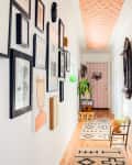 Hallway with framed art on walls, patterned ceiling, potted plants, and geometric rugs leading to a pink door.
