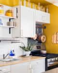 White kitchen cabinets, yellow wall, hexagonal backsplash, hanging pots, wooden countertop, and potted plant near stove.