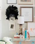 Gallery wall with framed art, a feathered mask, table lamp, books, and a vase with green foliage on a console table.