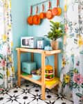 Kitchen corner with hanging orange pans, floral curtains, wooden cart with microwave, toaster, plant, and colorful decor.