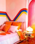 Colorful bedroom with pink bed, orange pillows, geometric wall art, and a gold side table with a lamp and books.
