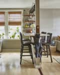 Open kitchen and living room with brown cabinets, bar stools, plants on windowsill, and a gray sofa with colorful cushions.