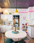 Colorful kitchen with white cabinets, round table, yellow pendant light, pink neon accents, and striped backsplash.