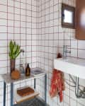 Bathroom with red grid tiles, round mirror, small window, white sink, plant, and toiletries on a marble shelf.