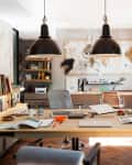 Home office with large desk, books, art supplies, and modern decor. Two pendant lights hang above, and a plant sits by the window.