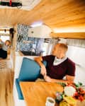 Man sitting in a cozy bus conversion with wood accents, blue seating, floral backsplash, and a child swinging from a door.