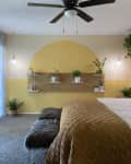 Bedroom with yellow accent wall, floating shelves, potted plants, brown quilted bedspread, and ceiling fan.