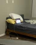 Unmade bed with gray sheets, laptop, and cardboard box in a sparse room.