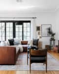 Living room with brown leather sectional, black-framed windows, wooden sideboard, and decorative plants.
