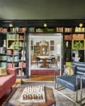 Living room with red sofa, blue armchair, chessboard, and bookshelves leading to a dining room with a chandelier.