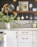 Kitchen with marble countertops, blue stove, hanging mugs, glassware, and a vase with greenery on a shelf.