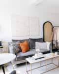 Studio apartment with a gray sofa, white armchair, bed, clothing rack, and modern decor.