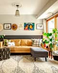 Living room with tan sofa, colorful pillows, wall art, large plants, and a patterned rug near sliding glass doors.