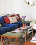 Cozy living room with a blue sofa, colorful cushions, glass coffee table, plants, and a round mirror on the wall.