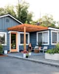 Blue house with orange door, wooden pergola, white chairs, potted plants, and raised garden beds in front yard.