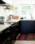 Modern kitchen with black cabinets, white countertops, red patterned rug, and hanging plants by a large window.