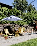 Patio with yellow chairs, striped umbrella, fire pit, and outdoor dining area next to a blue house with string lights.