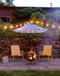 Patio with two yellow chairs, a striped umbrella, string lights, and a fire pit against a stone wall with ivy.