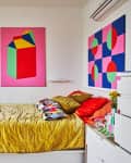 Bedroom with colorful abstract art, mustard bedspread, patterned pillows, and white furniture.
