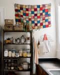 Laundry room with stacked washer and dryer, wooden shelves with jars, plants, and a colorful patchwork wall hanging.