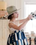 Woman in a patterned dress and hat arranging a geometric blanket in a bright room with stacked books.