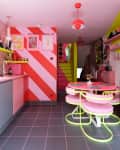 A midcentury kitchen with bright red and pink walls and cabinets, a small pink dining table, bright yellow wall shelves, and pink framed art prints on the wall
