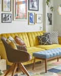Yellow sofa with colorful cushions, blue ottoman, gallery wall, and plants in a bright living room with large windows.