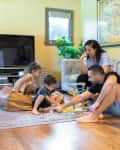 Family playing with building blocks on a living room floor, surrounded by a TV, plants, and a large map on the wall.