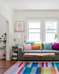 Living room with gray sofa, colorful pillows, wooden coffee table, plants, and a rainbow-striped rug.