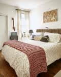 Cozy bedroom with a patterned bedspread, pink knit throw, guitar on wall, and bedside table with lamps and decor.