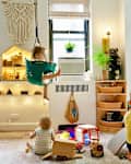 Children playing in a colorful playroom with toys, a hanging swing, and a dollhouse under a macrame wall hanging.