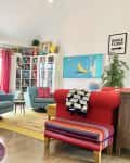Colorful living room with blue sofas, red armchair, large plant, and vibrant decor, including a banana painting and glass table.