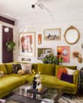 Colorful living room with green sectional sofa, orange curtains, eclectic wall art, and various plants.