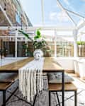 Sunlit conservatory with wooden dining table, bench seating, white runner, and potted plants, surrounded by glass walls.
