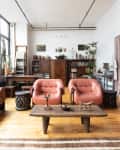 Loft living room with large windows, leather sofas, wooden coffee table, potted plants, and eclectic decor.