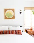 Bedroom with white bedding, colorful striped pillow, wooden nightstands, wall art, and potted plant by the window.