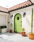 Spanish-style entryway with a green arched door, terracotta roof, potted plants, and patterned tile floor.