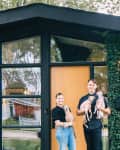 Couple holding dogs in front of a modern black house with ivy-covered walls and large windows.