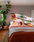 Bedroom with large plants by a window, orange bedding, colorful pillows, and framed art above the bed.
